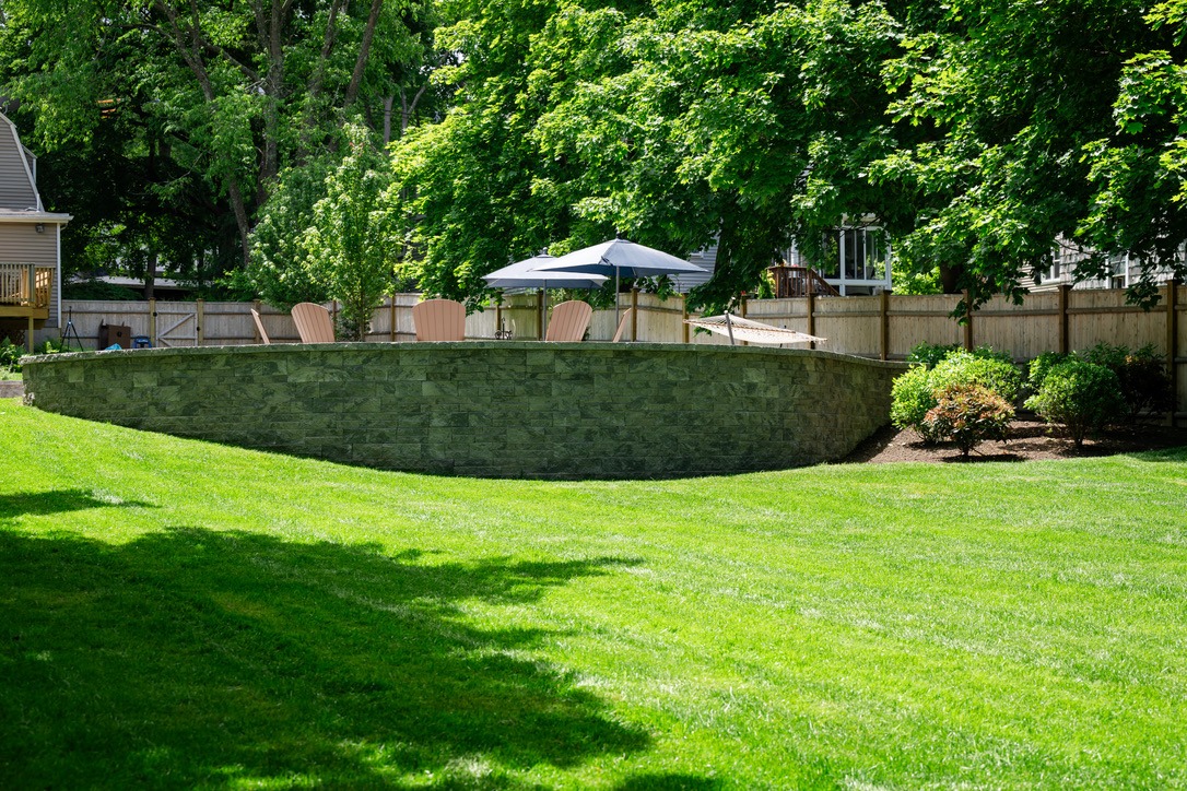 A lush backyard with a stone wall, Adirondack chairs, and an umbrella, surrounded by trees and fenced wooden privacy panels.