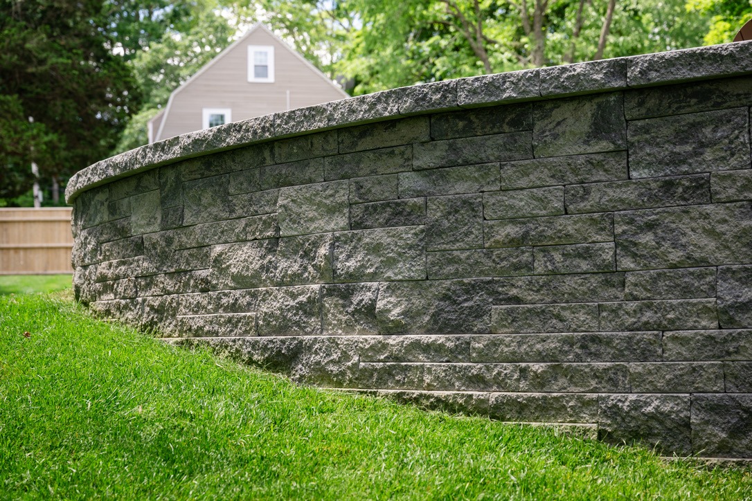 Curved stone retaining wall with vibrant green grass, a wooden fence, and a house in the background, surrounded by lush trees.