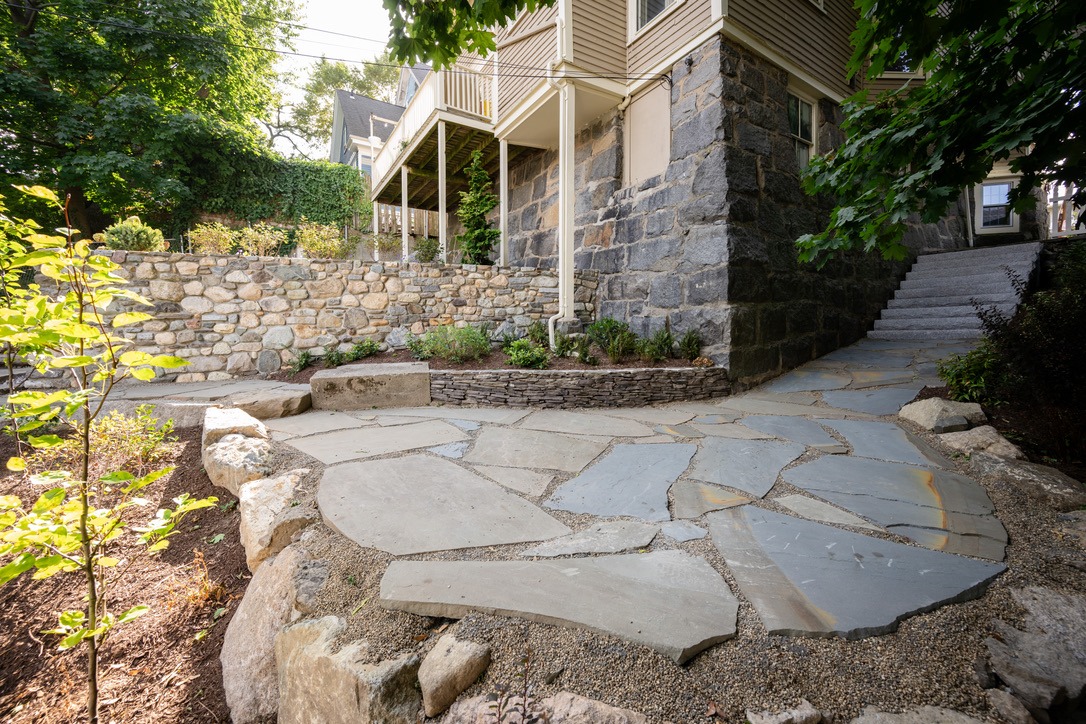 Stone house with a raised porch, surrounded by a rustic stone wall and garden, situated on a paved stone pathway under sunny foliage.