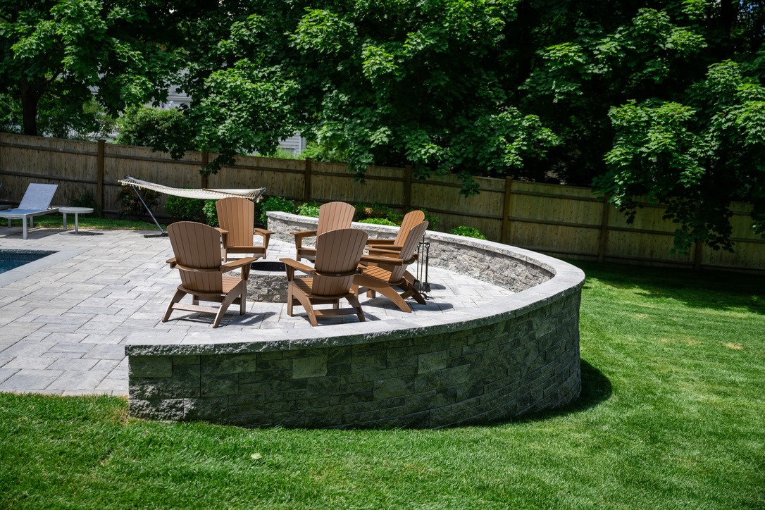 A patio area with wooden chairs surrounds a fire pit. Nearby is a hammock next to a grassy yard and wooden fence.