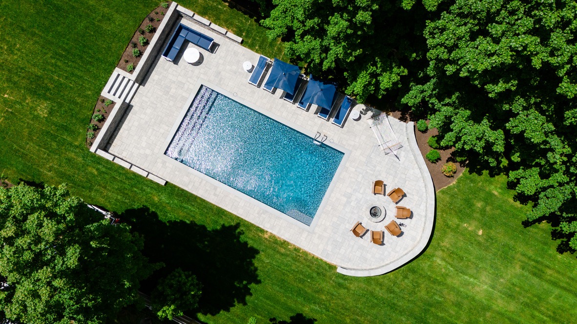 Aerial view of a modern rectangular pool with sun loungers, patio furniture, and surrounding greenery in a spacious outdoor setting.