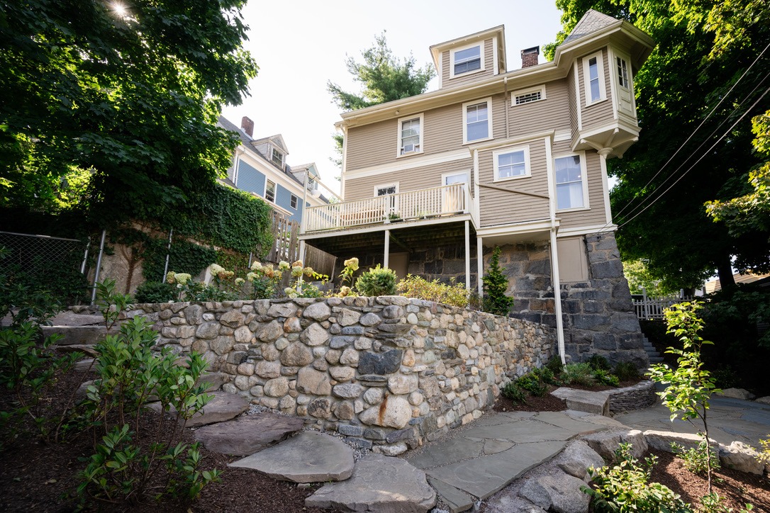 A large, historic wooden house with a stone foundation, surrounded by trees and a stone wall, under a clear blue sky.