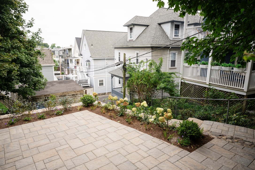 A paved patio with flower beds overlooks a residential area of gray houses and lush greenery, creating a serene suburban scene.