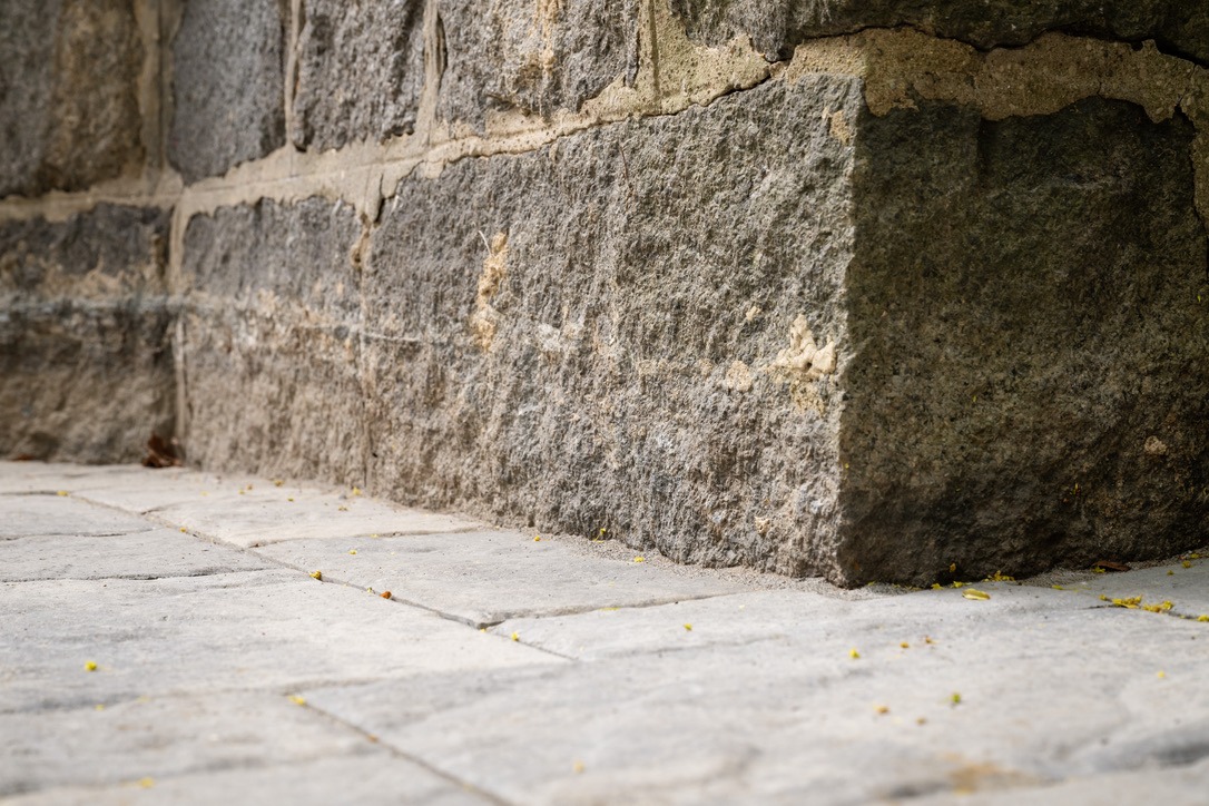 Close-up of a stone wall corner and paved ground, with small yellow flowers scattered. Texture and details of masonry are evident.