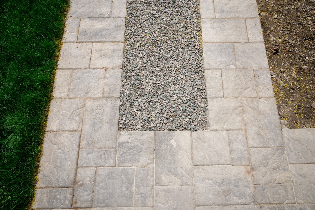 Stone pathway with gravel center, bordered by green grass and exposed soil. The geometric design creates a modern, clean outdoor aesthetic.