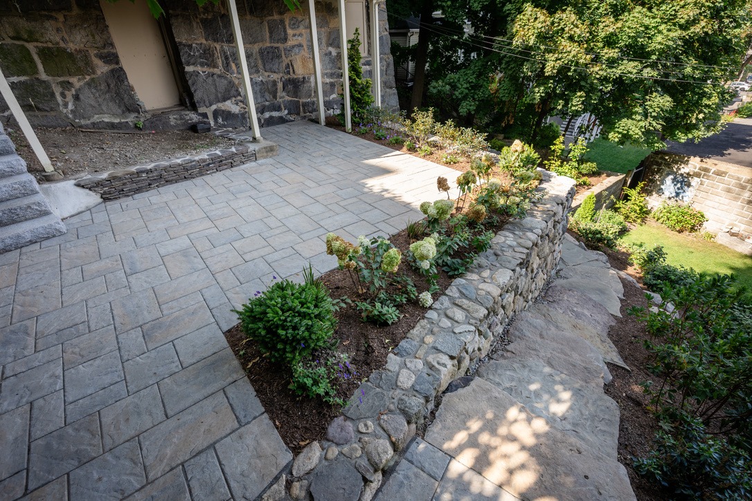 Stone patio with landscaped garden, paver walkway, and stone retaining wall. Green trees and grass visible in the sunny background.