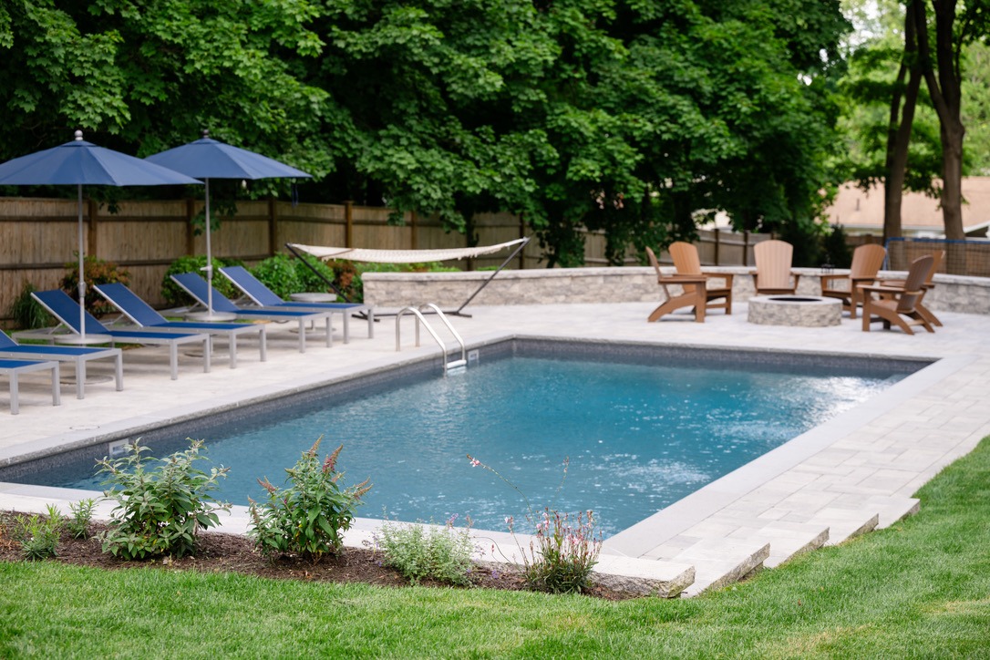 A serene backyard scene with a pool, lounge chairs, umbrellas, and a hammock, surrounded by lush greenery and a wooden fence.