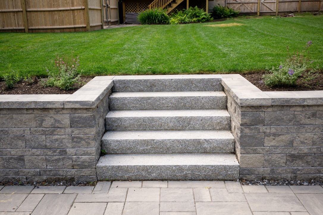 Stone steps lead to a green lawn, bordered by flowerbeds and wooden fencing. Neatly maintained garden creates a peaceful outdoor space.