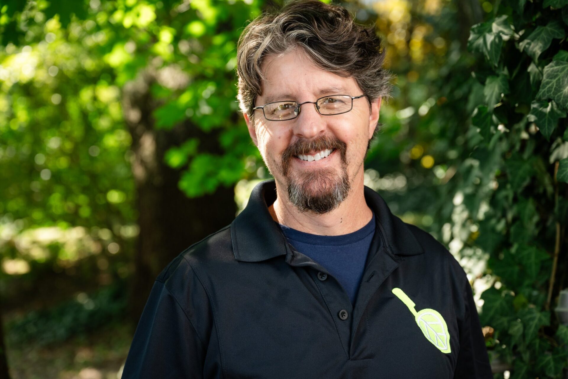 A person wearing glasses and a black shirt with a green leaf emblem smiles outdoors, standing against a leafy, green background.