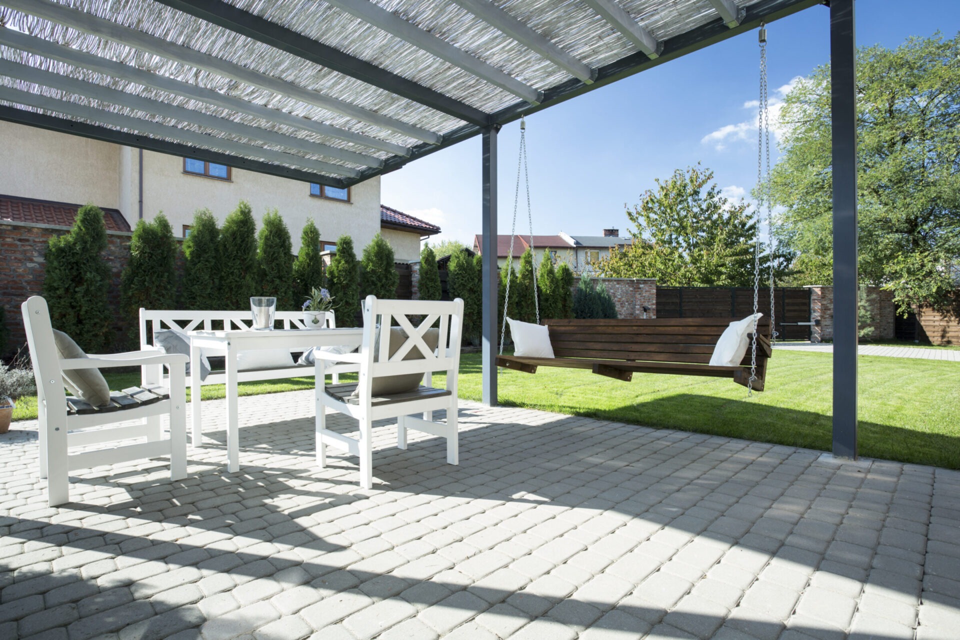 A tranquil patio with white furniture, wooden swing, and lush green garden under a pergola. Houses and trees in the background.